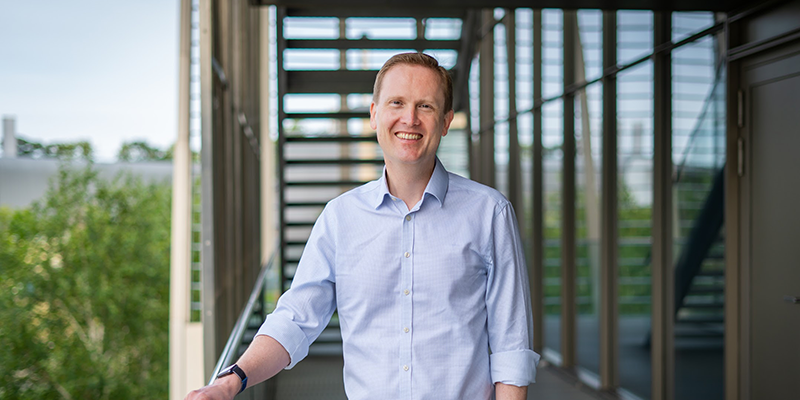 A man in a blue shirt is standing on the balcony of an office building made of metal and glass. He is posing and smiling at the camera, left hand in his pocket and right hand resting on the balcony railing. Surrounding trees can be seen in the background and reflected in the building.